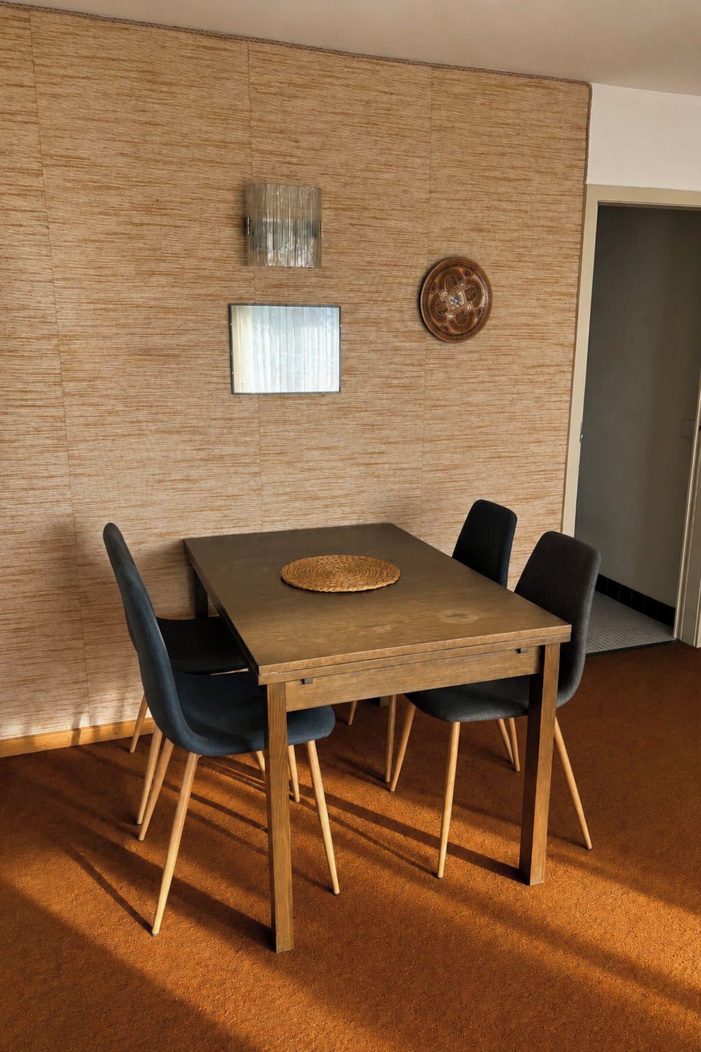 A dining area with a wooden table and four chairs, a woven placemat, and a textured wall with a decorative plate and a framed mirror.