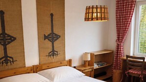 Una cama de madera con sábanas blancas y una pila de toallas, un tapiz de pared tejido, una lámpara colgante y una cortina a cuadros.