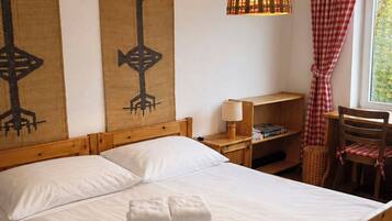 A wooden bed with white linens and a stack of towels, a woven wall hanging, a hanging lamp, and a checkered curtain.