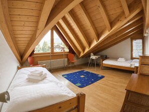 A dormitory room with a sloped ceiling, wooden beams, a bed, a desk, a chair, a radiator, and a window with a view of trees. - Residenz Darianne Leukerbad # 4-bed apartment, Dusche, WC by Interhome (Leukerbad)