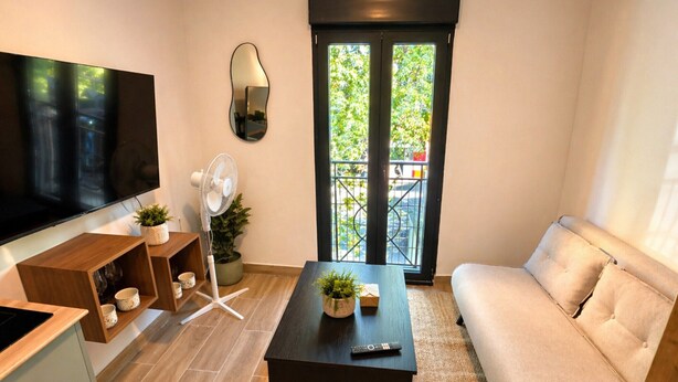 A modern living room with a flat-screen TV, a standing fan, a sofa, a coffee table, and a sliding glass door leading to a balcony. - Chic Madrid Stay With Terrace (Madrid)