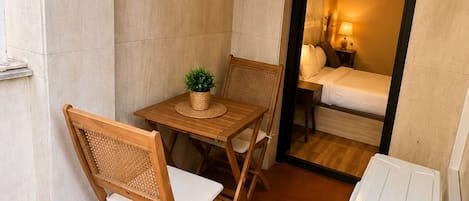 Ein kleiner Holztisch mit zwei Stühlen, ein Blumentopf und Blick in ein Schlafzimmer mit Bett und Nachttisch.