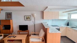 Kompakti oleskelutila, jossa on sohva, sohvapöytä, televisio, lattiavalaisin ja pieni keittiö.