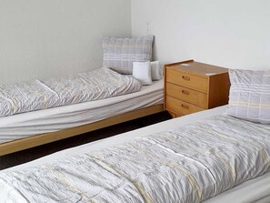 Een eenvoudige kamer met twee eenpersoonsbedden, een houten dressoir met laden en een effen wand.