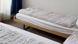 Yhden hengen sänky, jossa on puinen runko ja valkoiset vuodevaatteet. Huoneessa on pelkistetty valkoinen seinä ja ikkuna, jossa on siniset verhot.