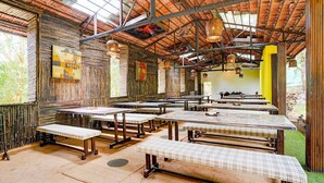 A rustic dining area with wooden tables and benches, a high ceiling with exposed beams, and a view of greenery outside.