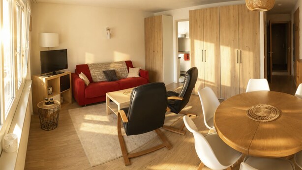 Een moderne woonkamer met een rode bank, een televisie, een eethoek met een ronde houten tafel en een keuken op de achtergrond.