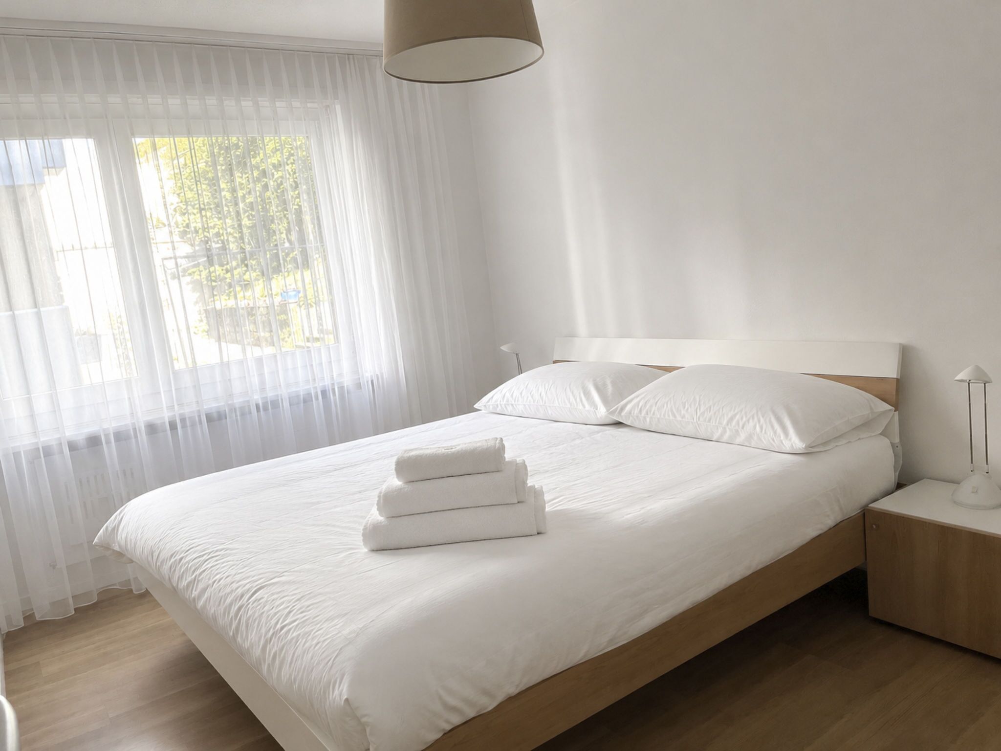 A neatly made bed with white linens and pillows, a bedside lamp, and a window with sheer curtains.