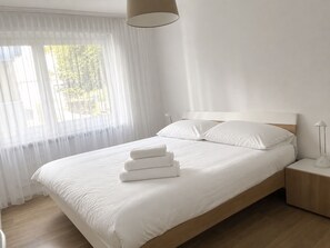 A neatly made bed with white linens and pillows, a bedside lamp, and a window with sheer curtains. - Majesta Leukerbad # 6-bed apartment, Dusche und Bad, WC by Interhome (Leukerbad)