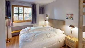 Et soveværelse med to senge, et vindue med gardiner, et natbord med en lampe og et billede hængende på væggen.