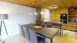 A modern dining area with a wooden table and chairs, a built-in kitchen, and a ceiling with exposed beams. - Apartment Skyline 406 by Interhome (St. Moritz)