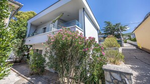 Une maison moderne à deux étages, dotée d’un balcon et entourée de verdure et de plantes en fleurs.