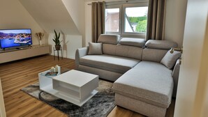 Un salon moderne avec un canapé d’angle, un téléviseur à écran plat, une table basse et une fenêtre avec des rideaux.