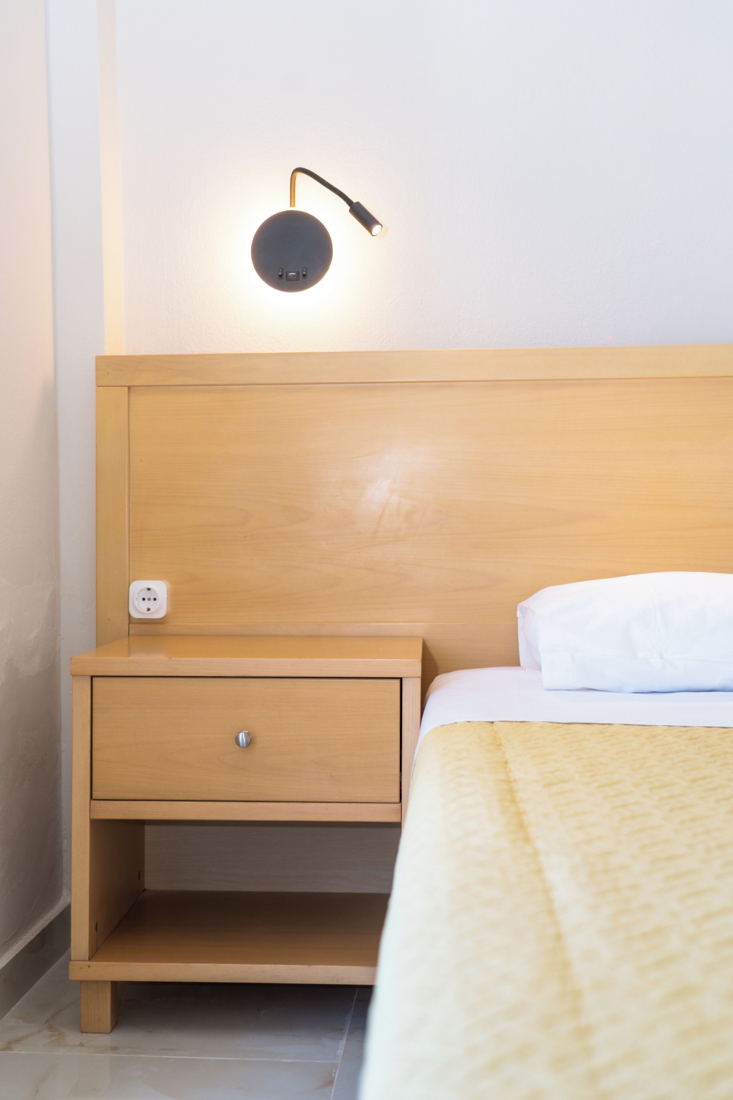 Una cama bien hecha con un cojín blanco y una mesita de noche de madera con cajón y lámpara.