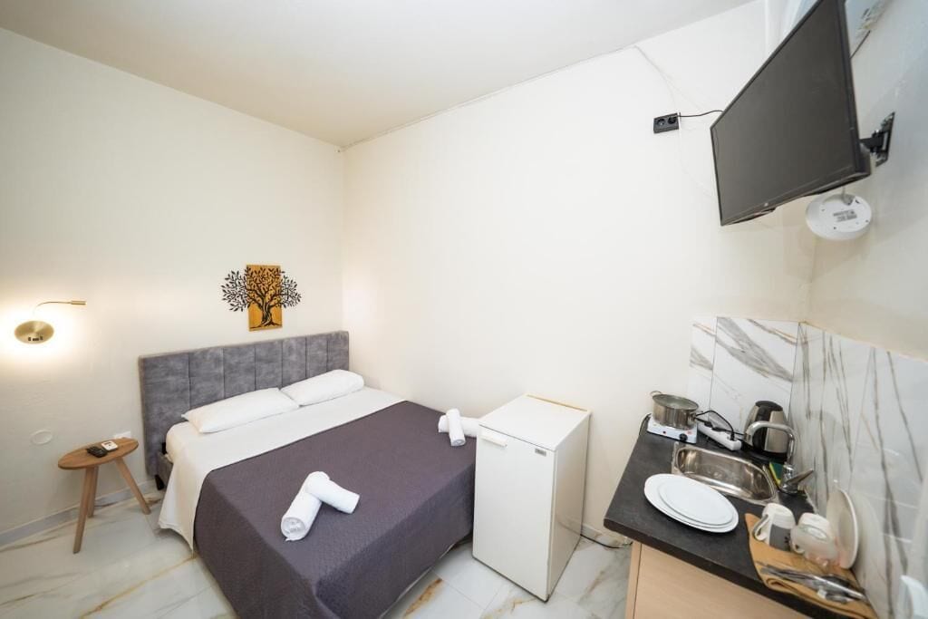Et hotelværelse med en seng, et lille skrivebord med håndvask, et fjernsyn ophængt på væggen og et lille bord med en lampe.