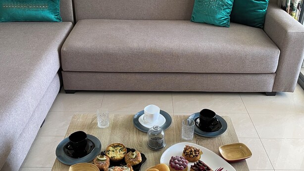 A living room with a grey sofa, teal cushions, and a coffee table with various food items and drinks. - Design Apt @ Blanca Beach: Private Beach & Pool (Sidi Rahal)