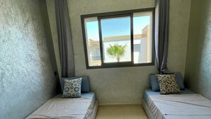 A room with two beds, a window with curtains, a view of buildings, and a tiled floor. - Design Apt @ Blanca Beach: Private Beach & Pool (Sidi Rahal)
