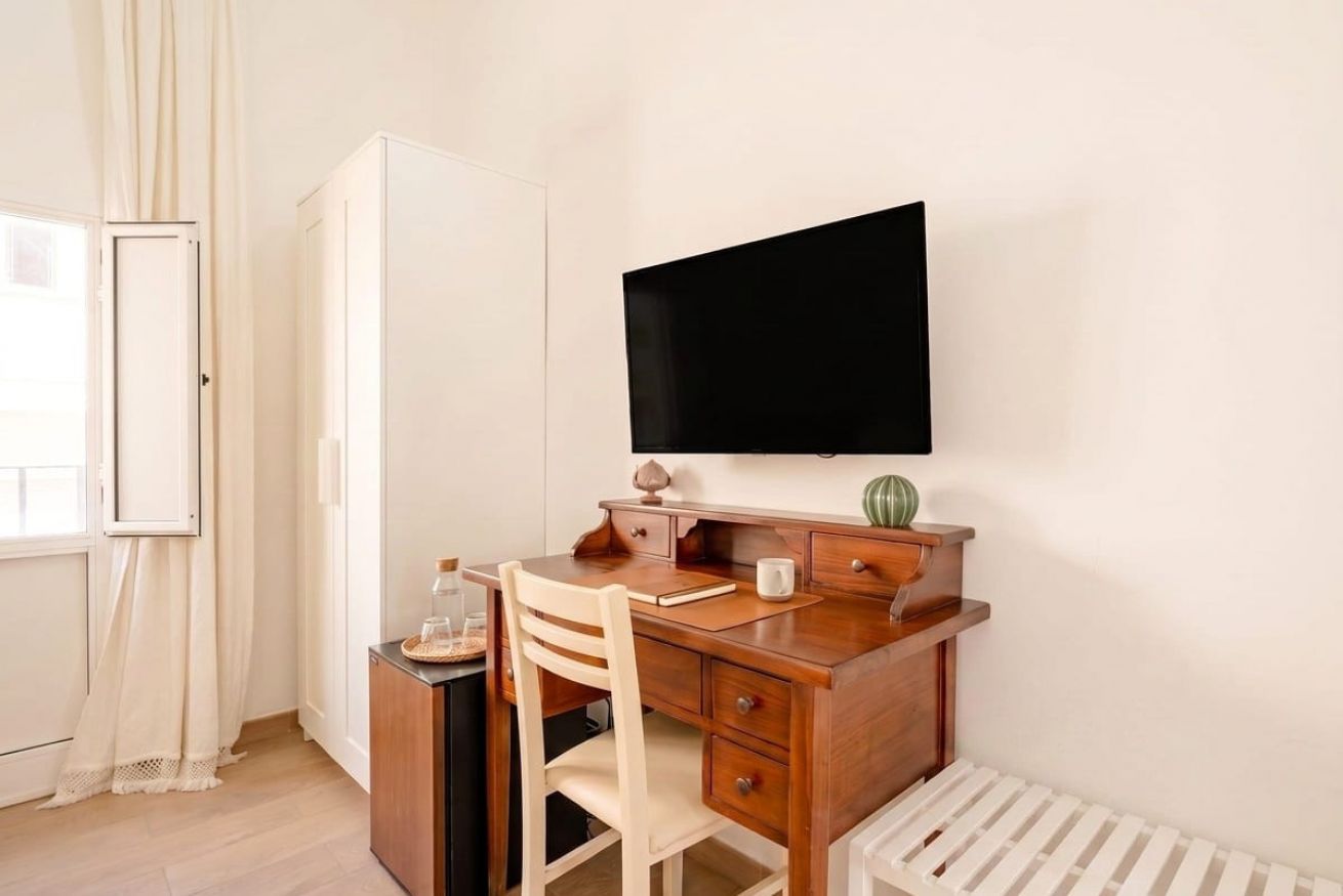 Ein Zimmer mit Schreibtisch aus Holz, einem Sessel, einem an der Wand montierten Fernseher, einem Heizkörper und einem Kleiderschrank.