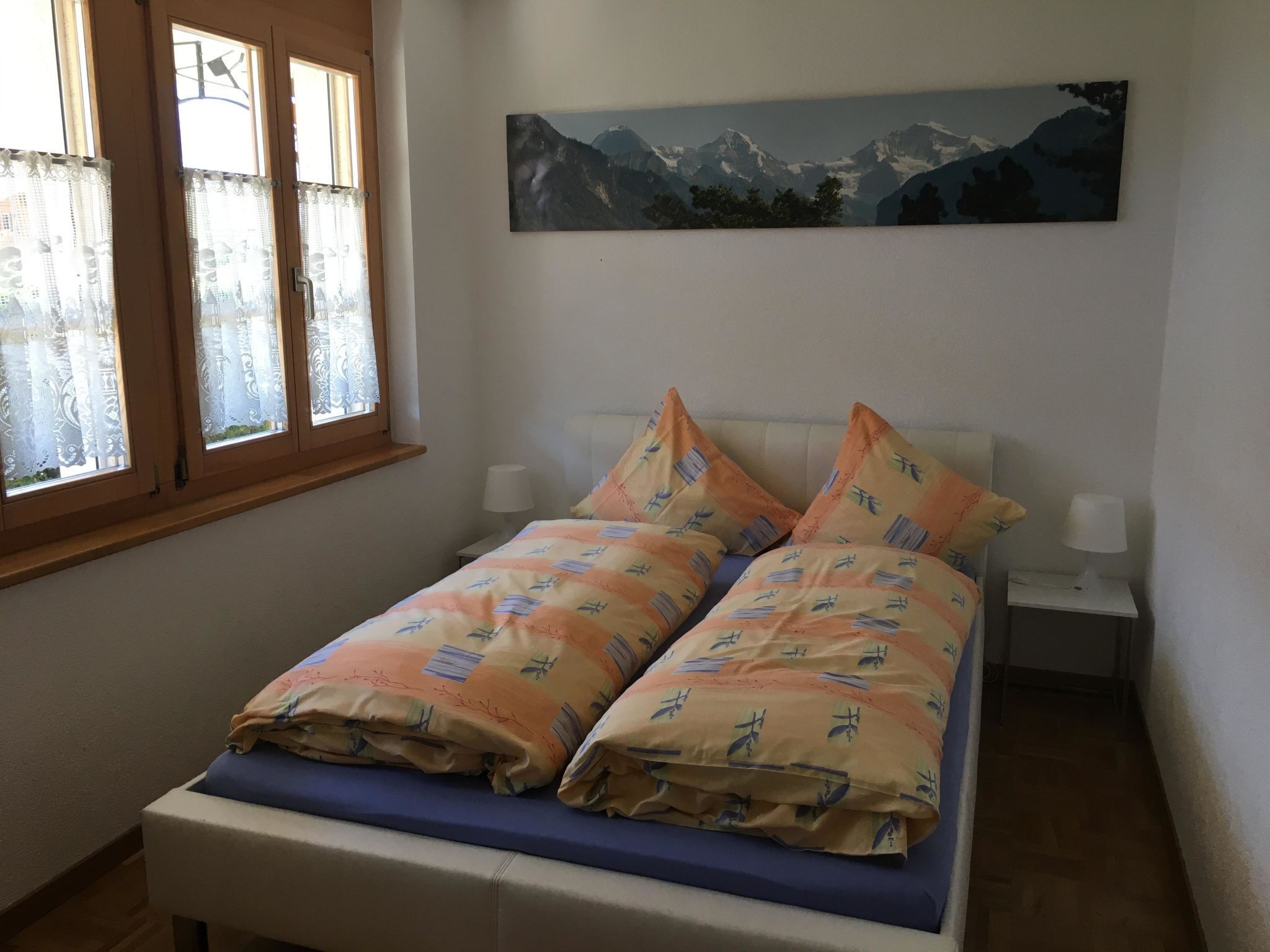 A bedroom with a bed, a window with curtains, a nightstand, and a painting on the wall.
