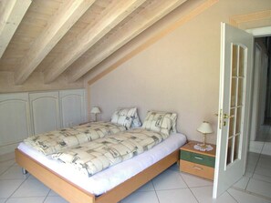 A bedroom with a wooden ceiling, a bed with patterned bedding, a nightstand with a lamp, and a door leading to another room. - Adlerhorst Leukerbad # 6-bed apartment, Dusche oder Bad, WC by Interhome (Leukerbad)