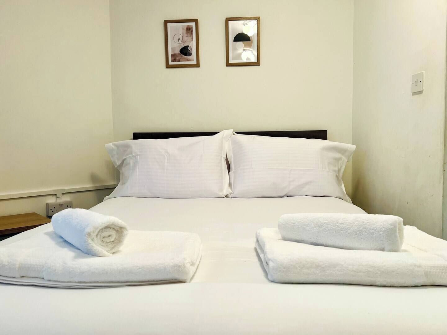 A hotel room with a bed, two pillows, two bedside tables with towels, and two framed pictures on the wall.