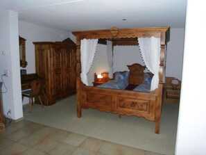 Un lit à baldaquin, une table de chevet en bois avec une lampe et une chaise dans une chambre dont le sol est recouvert de carreaux.
