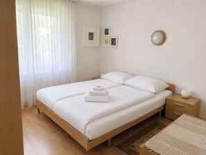 A neatly made double bed with white linens and two folded towels, a bedside table with a lamp, and a small rug on the floor. - Majesta Leukerbad # 6-bed apartment, Dusche und Bad, WC by Interhome (Leukerbad)