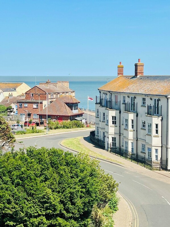 A coastal town with a prominent white building with multiple windows and a flag, a curved road, and a clear view of the sea.
