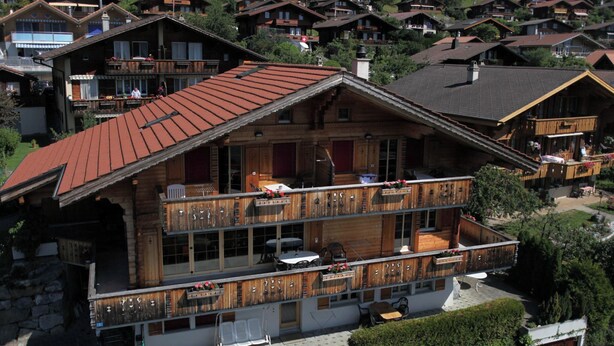 Een meerlaags houten chalet met balkons, een stenen fundering en een rode dakbedekking.