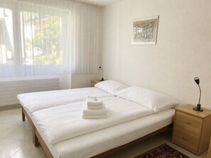 A hotel room with a double bed, a bedside table with a lamp, a small drawer, a window with sheer curtains, and a wall-mounted picture. - Majesta Leukerbad # 6-bed apartment, Dusche oder Bad, WC by Interhome (Leukerbad)