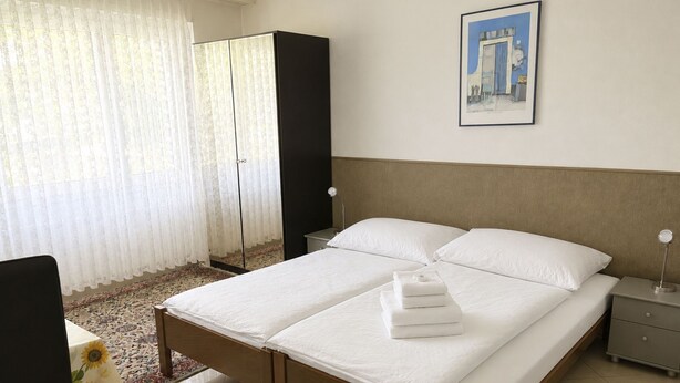 A hotel room with a double bed, a bedside table, a nightstand, a wardrobe, a window with sheer curtains, and a framed picture on the wall. - Majesta Leukerbad # 6-bed apartment, Dusche oder Bad, WC by Interhome (Leukerbad)