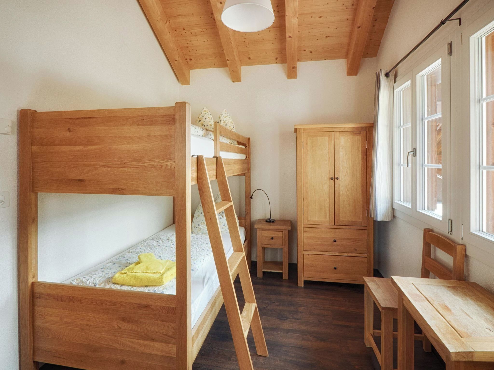 Una cama litera con escalera, un armario de madera, una mesita y una ventana con cortinas.