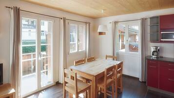A dining area with a wooden table and chairs, a kitchen with red cabinets, and large windows with curtains.