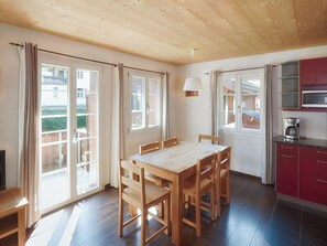 Un comedor con una mesa y sillas de madera, una cocina con armarios rojos y ventanas grandes con cortinas.