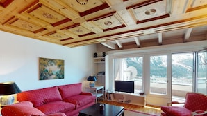 A living room with a red sofa, a black coffee table, a red armchair, and a television. There is a painting on the wall and a window with a view of mountains.