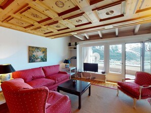 A living room with a red sofa, a black coffee table, a red armchair, and a television. There is a painting on the wall and a window with a view of mountains. - Apartment Tinus by Interhome (St. Moritz)