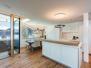 A modern kitchen with a wooden floor, white cabinets, a dining area with a table and chairs, and a view of snow-capped mountains through large windows. - Apartment Mira Lej by Interhome (St. Moritz)