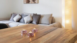 Une table en bois sur laquelle sont posés trois chandeliers roses, un canapé gris agrémenté de plusieurs coussins et, au mur, un tableau représentant une vache.