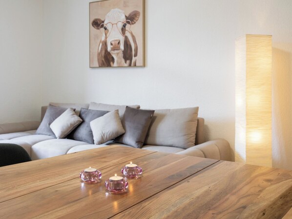 Ein Holztisch mit drei rosafarbenen Kerzenständern, ein graues Sofa mit vielen Kissen und ein Wandbild einer Kuh.