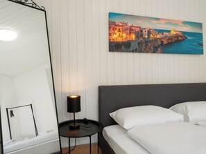 A bedroom with a bed, a bedside table with a lamp, a mirror, and a painting of a coastal town. - Apartment Troja Appartement/Fewo 4-Bettwohnung by Interhome (Interlaken)
