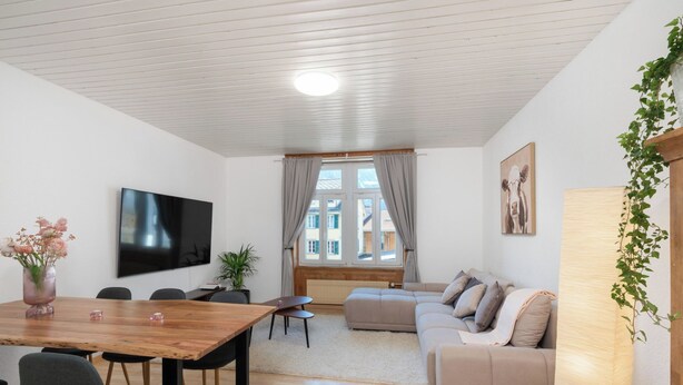 Un salon moderne avec une table à manger en bois, un canapé gris et un téléviseur fixé au mur.