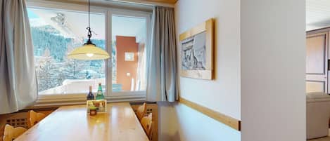 A dining area with a wooden table, chairs, a hanging light fixture, a window with curtains, and a framed picture on the wall.
