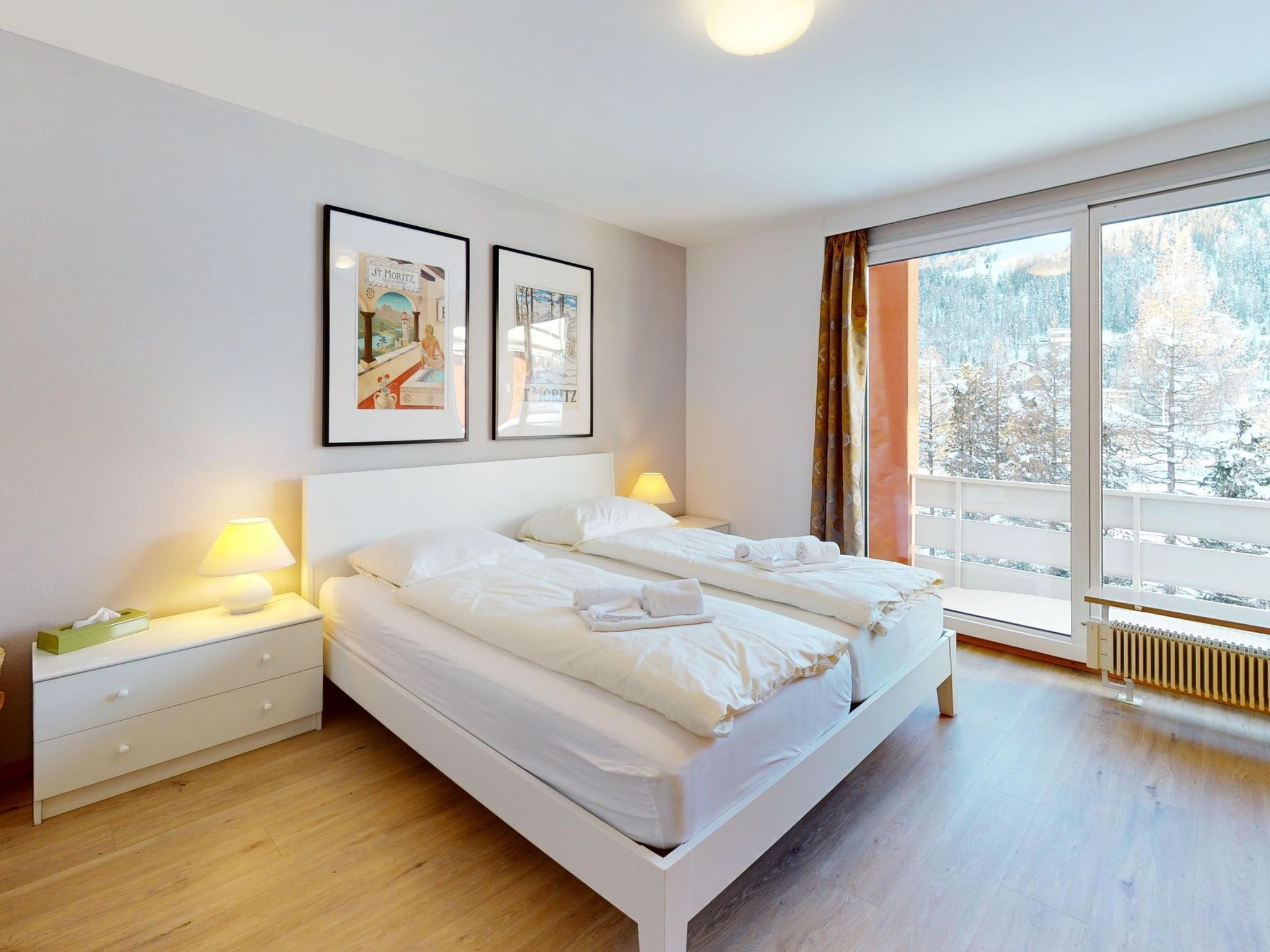 A hotel room with a large bed, a bedside table with a lamp, and a window with a view of snow-covered trees.