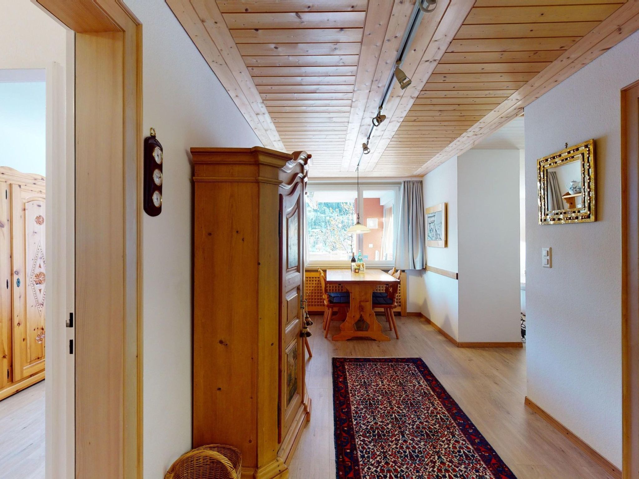 A hallway with wooden flooring, a dining area with a table and chairs, and a large wooden cabinet.