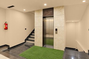 A modern elevator with a digital display, a fire extinguisher mounted on the wall, and a green artificial grass mat at the base of a staircase.