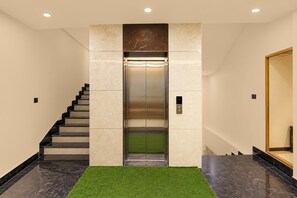Um elevador com visor digital, uma escada com degraus pretos e um tapete verde.