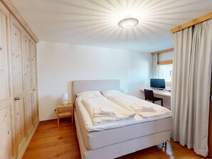 A bedroom with a bed, a desk, a chair, a wardrobe, and a window with curtains. - Apartment Arsa 28 by Interhome (Silvaplana-Surlej)