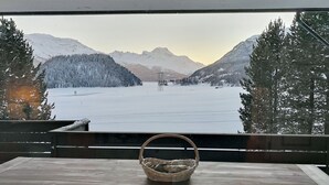 Een houten tafel met een gevlochten mandje voor een raam met uitzicht op een besneeuwd landschap met bergen.