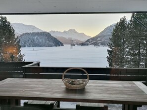 A wooden table with a woven basket in front of a window overlooking a snowy landscape with mountains. - Apartment Muntagna Alva OG by Interhome (St. Moritz)