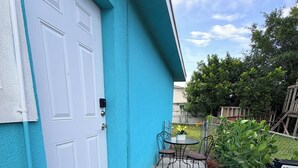 Uma casa azul com porta de garagem branca, um pequeno pátio com mesa e cadeiras, e um jardim com plantas e uma bomba d'água.